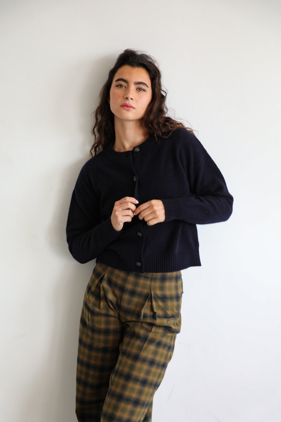 Model buttoning the Modern Buttons Knit Sweater Navy, a cropped 100% wool knit with button-down front, over the pleated grey and brown check Square Detail Pants.