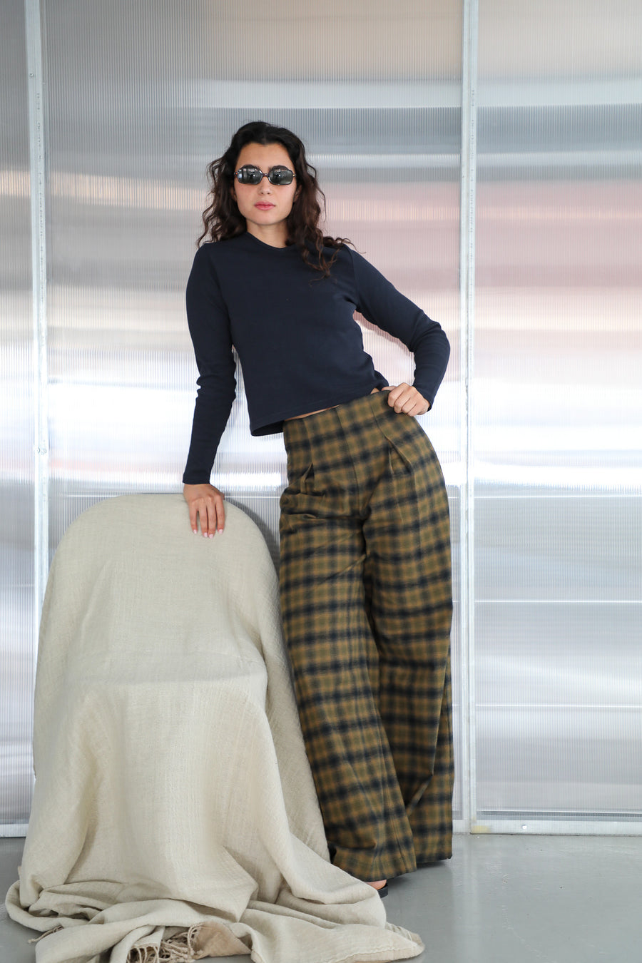 Model leaning on a chair in the Long V-Neck T-Shirt Navy, heavy organic cotton with long sleeves, tucked into the Square Detail Pants Grey, high-waisted wide-leg pleated trousers in a grey and brown check, 100% cotton.