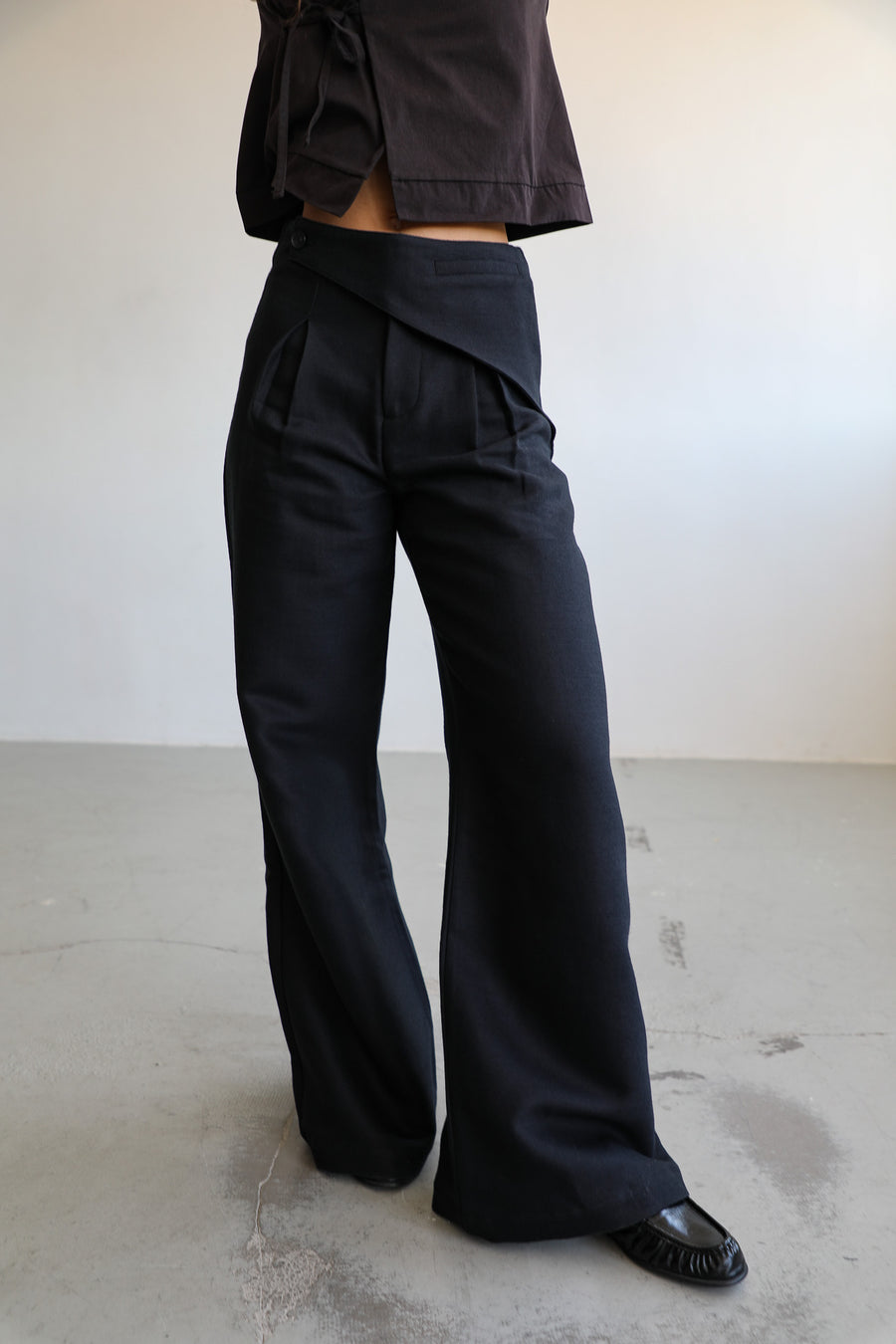 Close-up of the Wrap Pants Black: high-waisted wide-leg trousers with a crossed triangular waistband panel and front pleats, worn with the dark brown cotton Crossed Japanese Top.