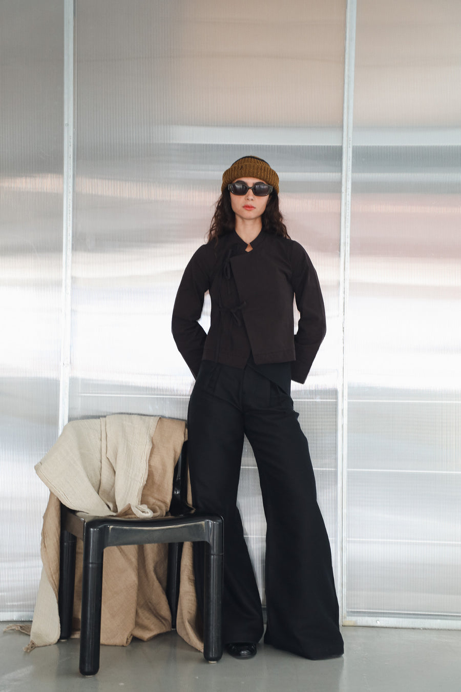 Model standing by a chair in the Crossed Japanese Top, dark brown 100% cotton with Perkins neckline and tied front, paired with the long wide-leg Wrap Pants Black.