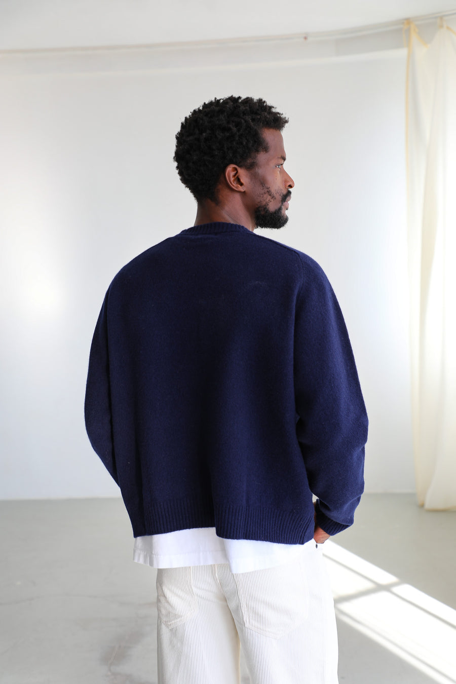 Back view of the Line Round Knit Sweater Dark Blue, a plain-back 100% wool knit with relaxed fit and ribbed hem, worn with the white wide-leg Corduroy Wide Comfy Pants in 100% cotton.