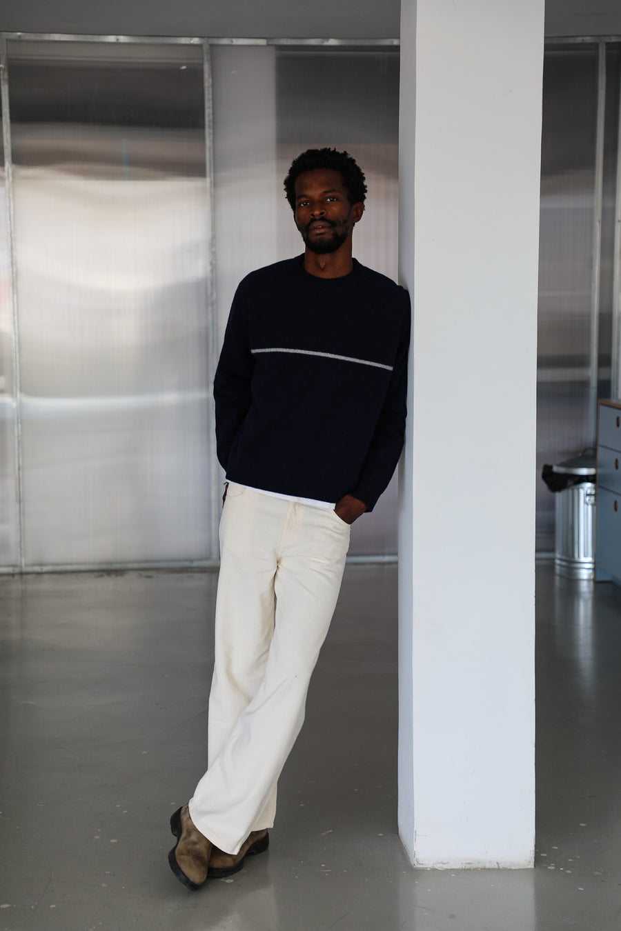 Model leaning on a column in the dark blue wool Line Round Knit Sweater with white chest line and the Corduroy Wide Comfy Pants White, long wide-leg trousers in 100% cotton corduroy.