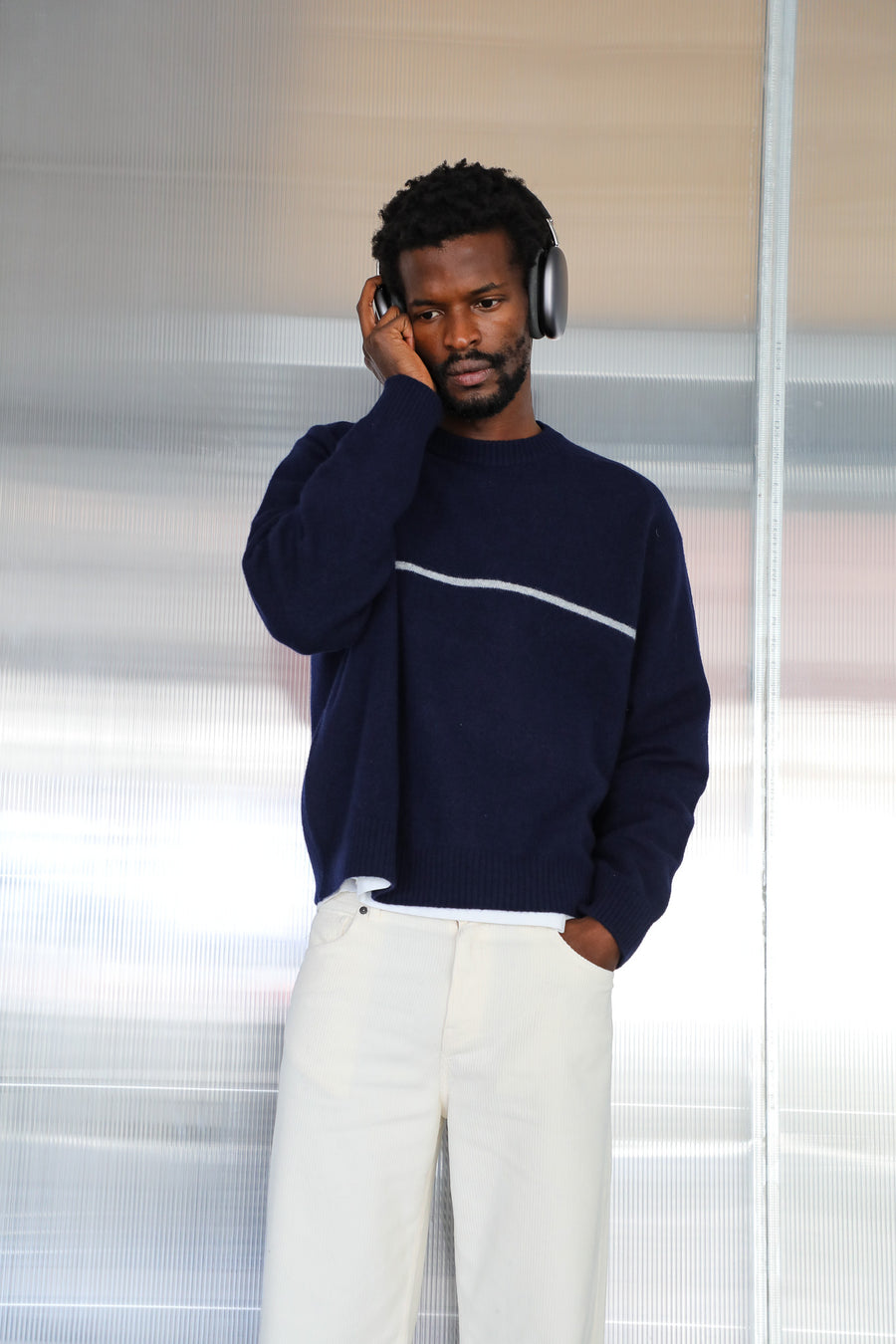 Model with headphones in the Line Round Knit Sweater Dark Blue, a relaxed 100% wool knit with round neck and white chest stripe, over the off-white wide-leg Corduroy Wide Comfy Pants in cotton.