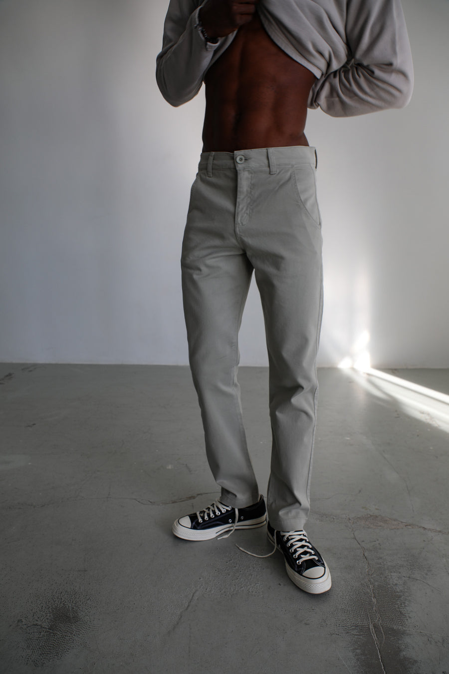 Lower-body view of the Button Comfy Pants Green: straight-leg sage green trousers in 100% cotton with slant side pockets and a mother-of-pearl front button, shown with the light grey cotton Polo Sweater lifted at the waist.