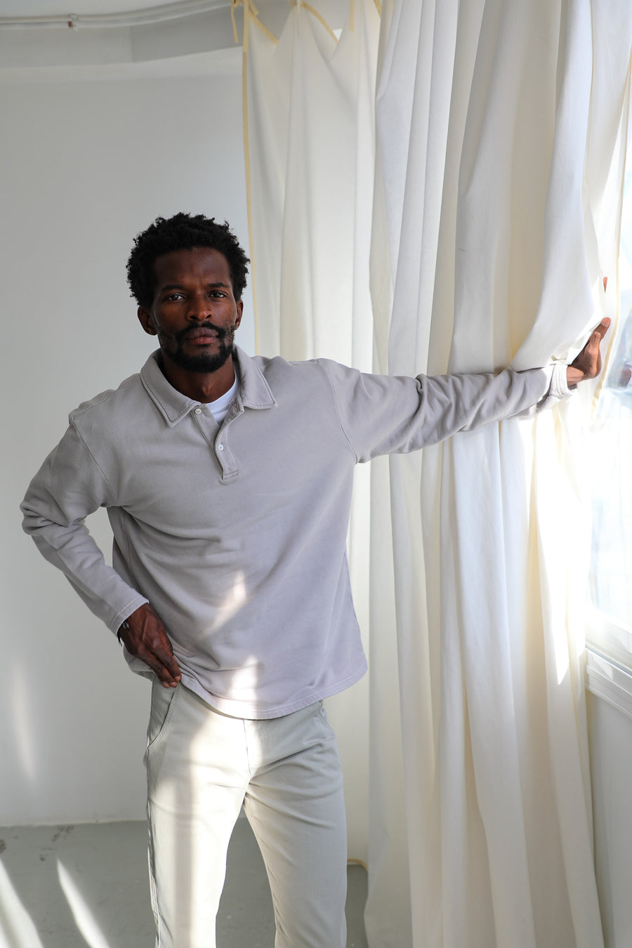 Model holding a white curtain in the Polo Sweater Light Grey, 100% cotton with soft polo collar, half-button placket and relaxed long sleeves, paired with the sage green 100% cotton Button Comfy Pants.