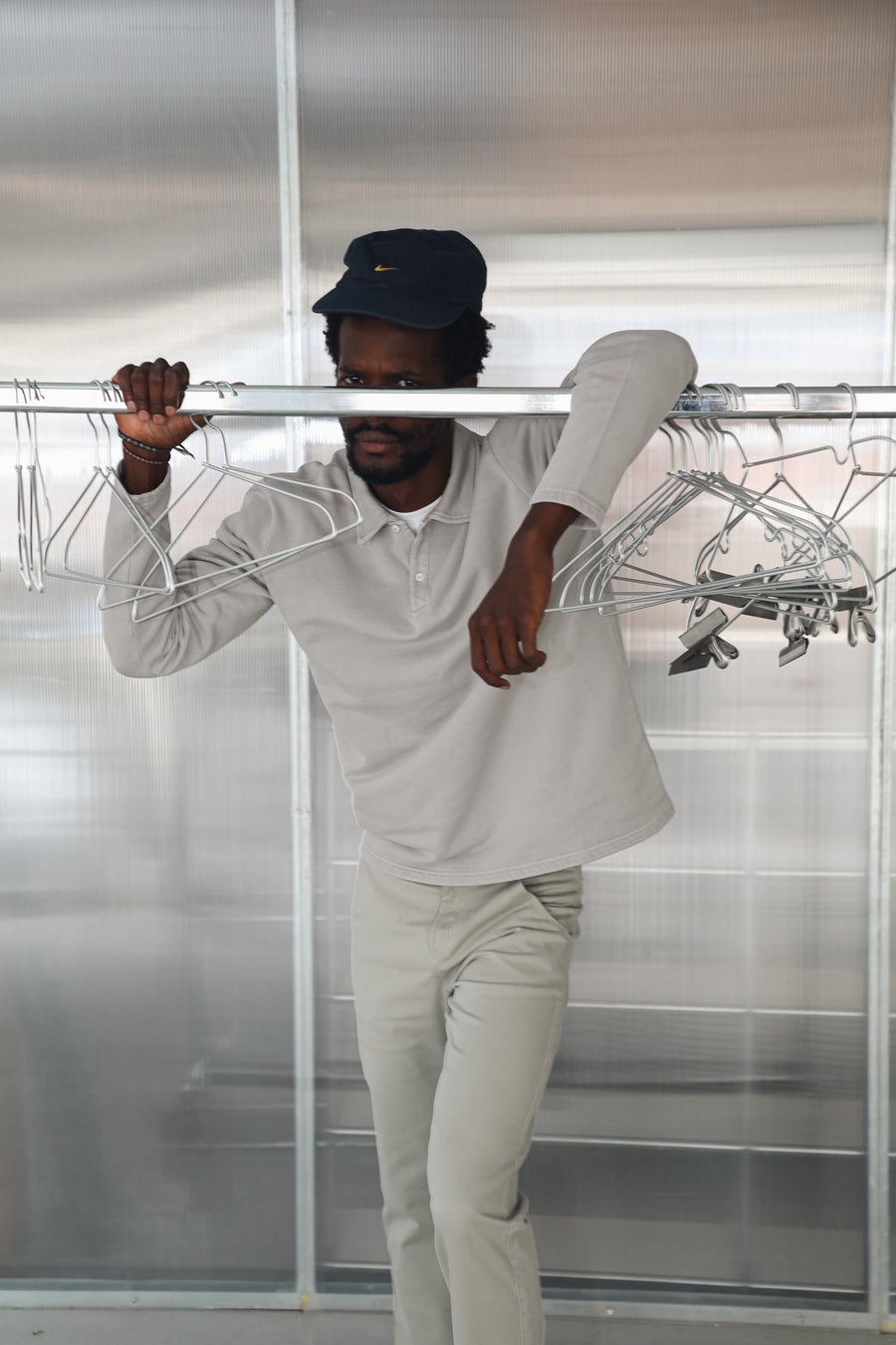 Model leaning on a clothing rail in the Polo Sweater Light Grey, 100% cotton with polo collar and buttoned placket, worn with the sage green 100% cotton Button Comfy Pants and a navy cap.